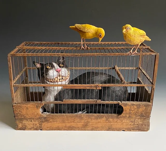 A ceramic cat in a birdcage, with two yellow birds perched above.